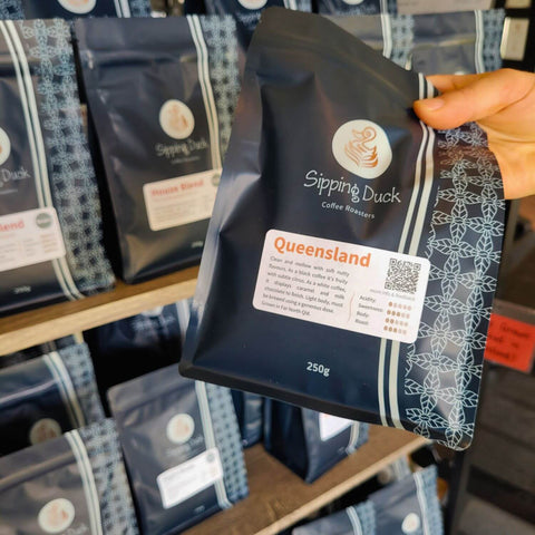Sipping Duck Coffee Roasters Queensland coffee in 250g bag with label describing the profile and coffee bags on a shelf in the background.