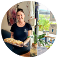 Emma smiling and holding a wooden board with almond croissants at Sipping Duck Straford. Plants and artwork.